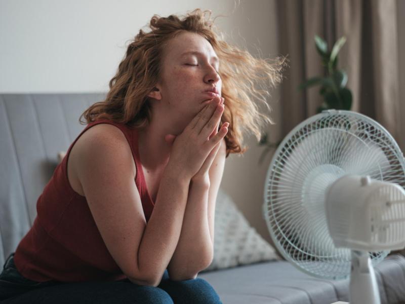 Frau sitzt auf der Couch und kühlt ihr Gesicht mit einem Ventilator.