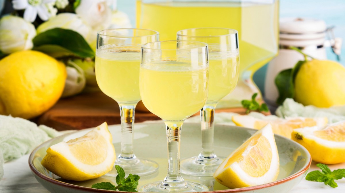 Drei kleine Gläser mit Limoncello stehen vor einer großen Flasche, umgeben von Zitronenspalten.