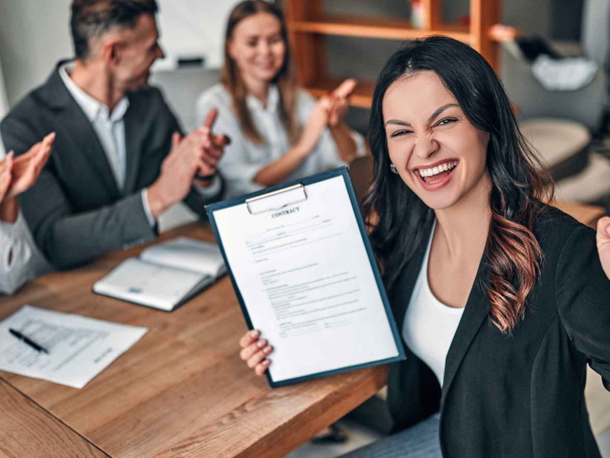 Junge Frau mit braunen Haaren sitzt auf der Arbeit und h&auml;lt einen Vertrag hoch, &uuml;ber den sie sich sehr freut.