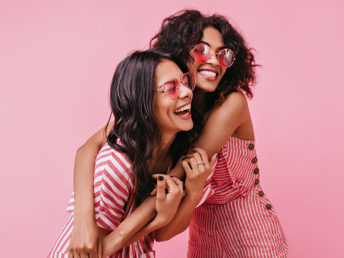 Zwei Frauen vor einer pinken Wand, die sich umarmen und lachen.