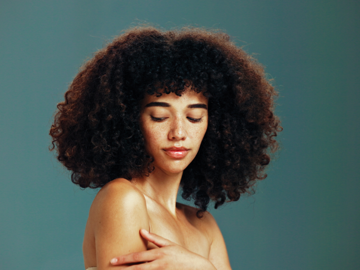 Junge Frau mit lockigem, dunklem Haar und Sommersprossen blickt mit geschlossenen Augen nachdenklich, vor schlichtem, grünlichem Hintergrund.