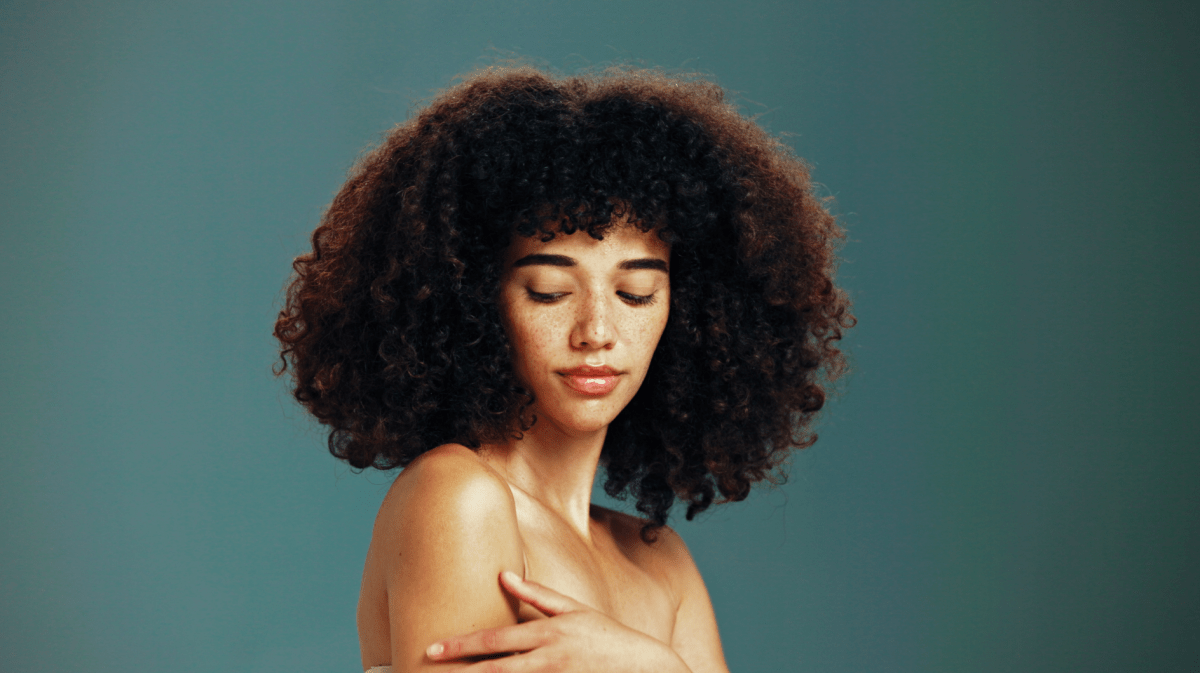 Junge Frau mit lockigem, dunklem Haar und Sommersprossen blickt mit geschlossenen Augen nachdenklich, vor schlichtem, grünlichem Hintergrund.