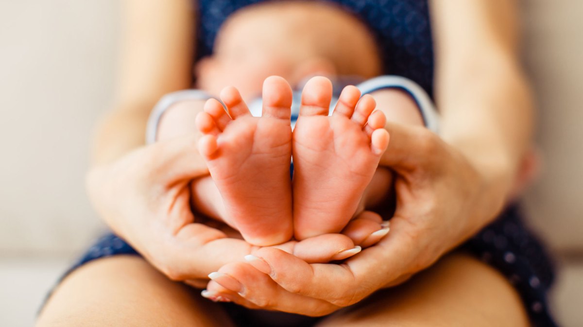 Nahaufnahme von Babyfüßen, die von den Händen der Mutter gehalten werden. Im Hintergrund erkennt man verschwommen, dass ein Baby auf dem Schoß seiner Mutter liegt.