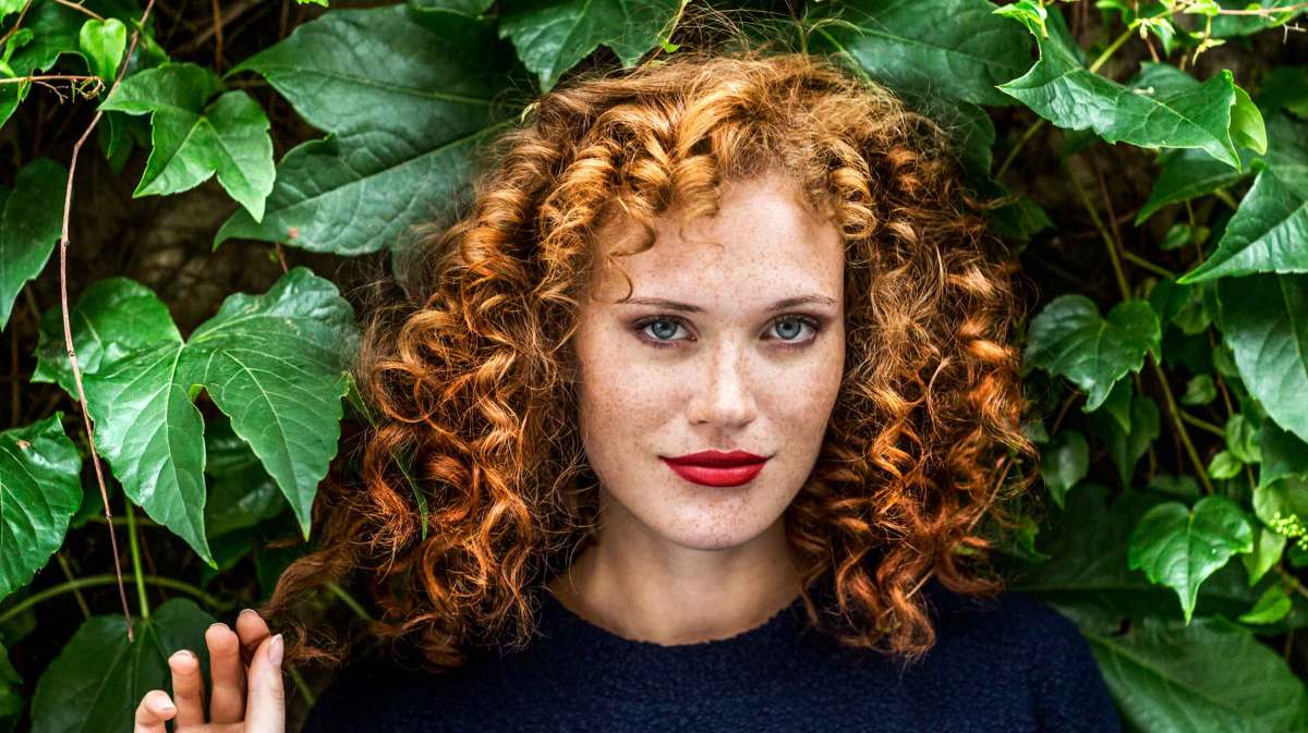 Junge Frau mit roten Locken, Sommersprossen und leuchtend roten Lippen blickt selbstbewusst vor einer grünen Blätterwand in die Kamera.