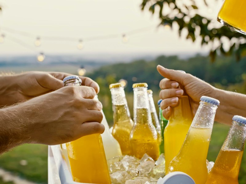 Mehrere Hände greifen nach gekühlten Flaschen mit gelbem Getränk aus einer mit Eis gefüllten Box im Freien, im Hintergrund Lichterketten und unscharfe Landschaft.