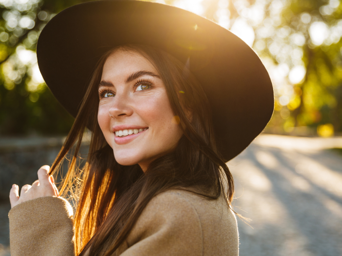 Frau im Sonnenlicht im Park, mit Hut auf dem Kopf, die sich umdreht und zur Kamera l&auml;chelt.