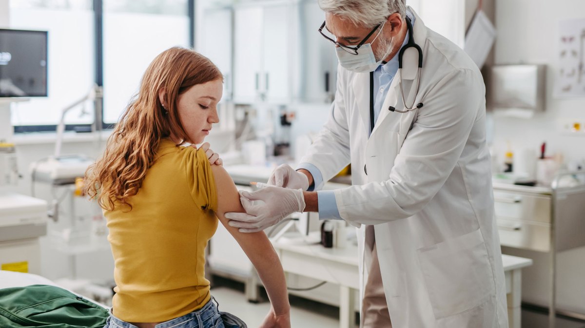 Mädchen, ca. 13 Jahre, sitzt in der Arztpraxis und erhält eine Spritze von einem älteren, männlichen Arzt.