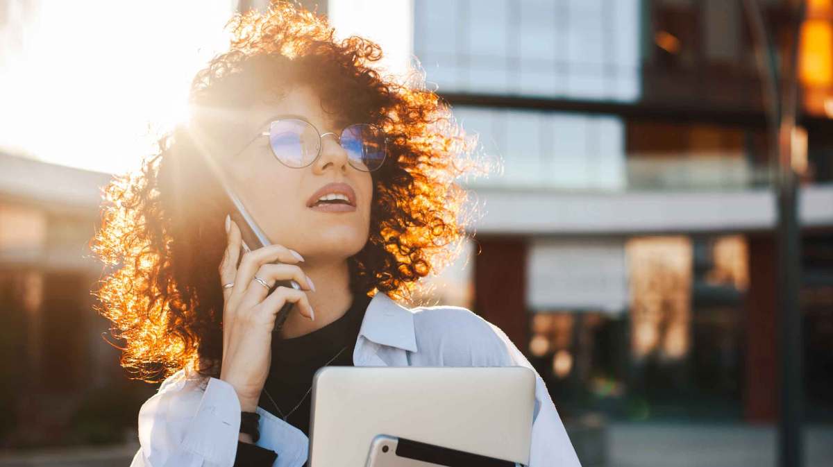 Junge Frau mit Brille und Locken telefoniert beruflich und hat ein Tablet im Arm.