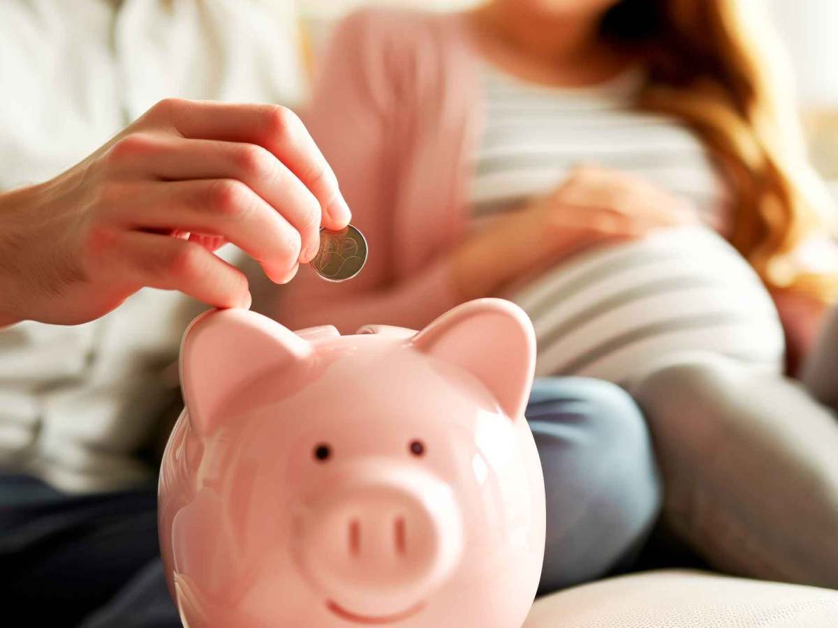 Schwangere Frau sitzt auf der Couch w&auml;hrend der Mann Geld in ein Sparschwein wirft.