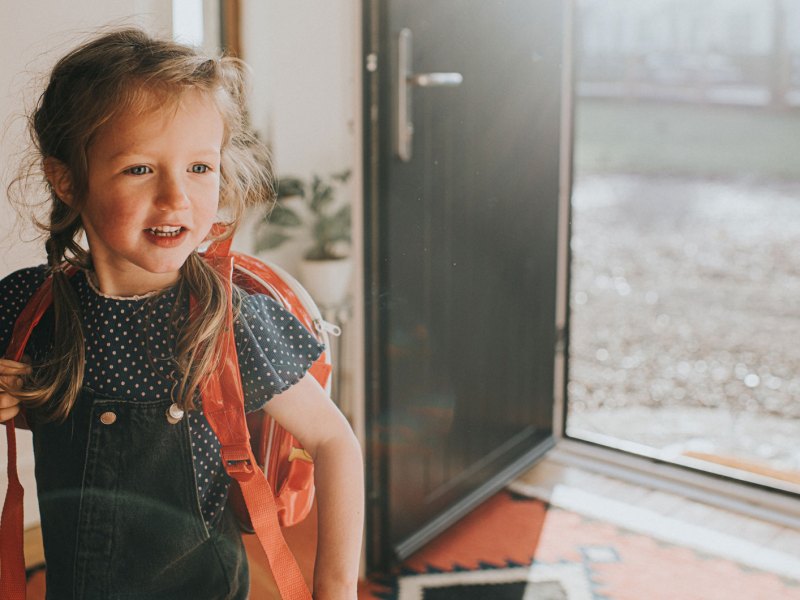 Mädchen mit Rucksack steht an einem Sommertag vor einer geöffneten Tür, bereit, loszugehen.