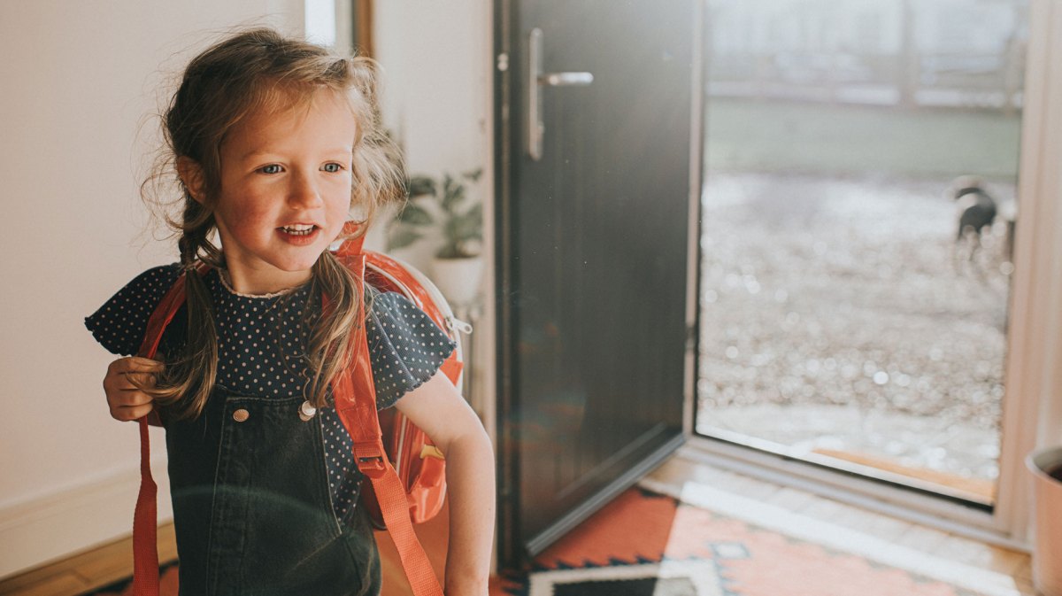 Mädchen mit Rucksack steht an einem Sommertag vor einer geöffneten Tür, bereit, loszugehen.