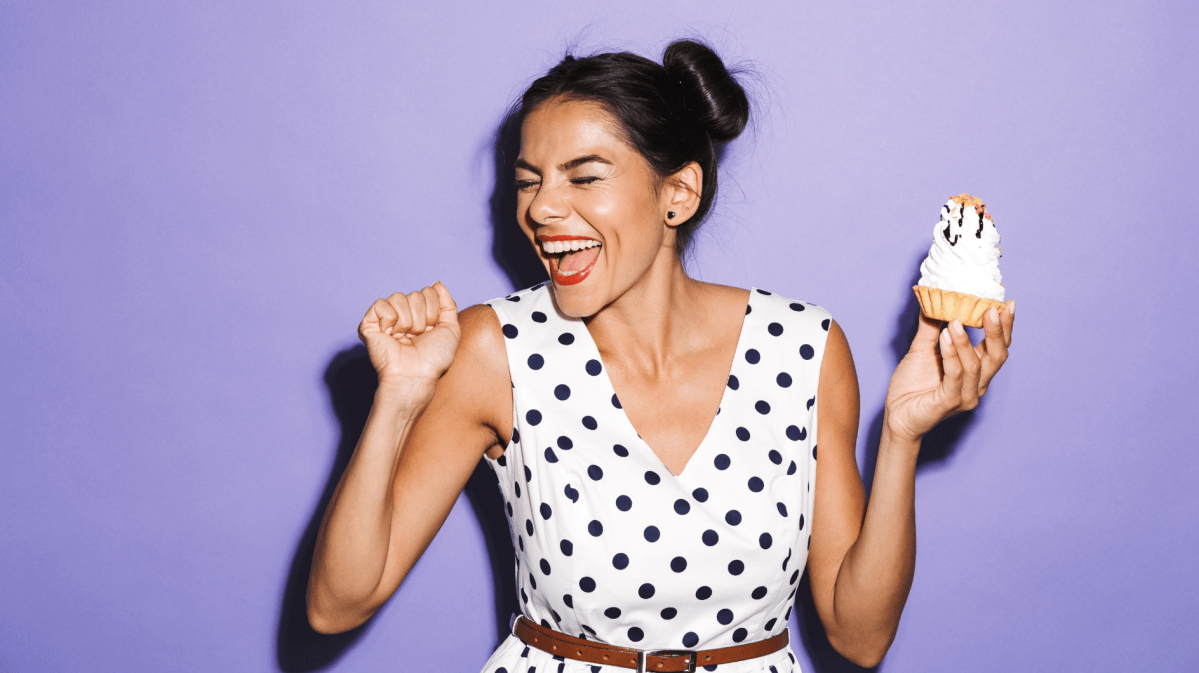 Frau vor einer lila Wand, mit einem Cupcake in der Hand, die tanzt.