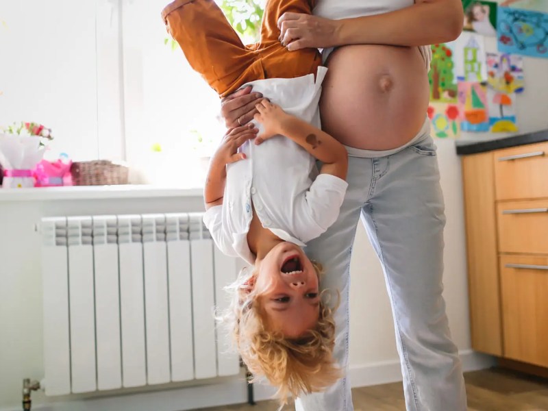 Schwangere Frau hebt ihr erstes Kind an den F&uuml;&szlig;en nach oben, das lacht herzlich dabei.