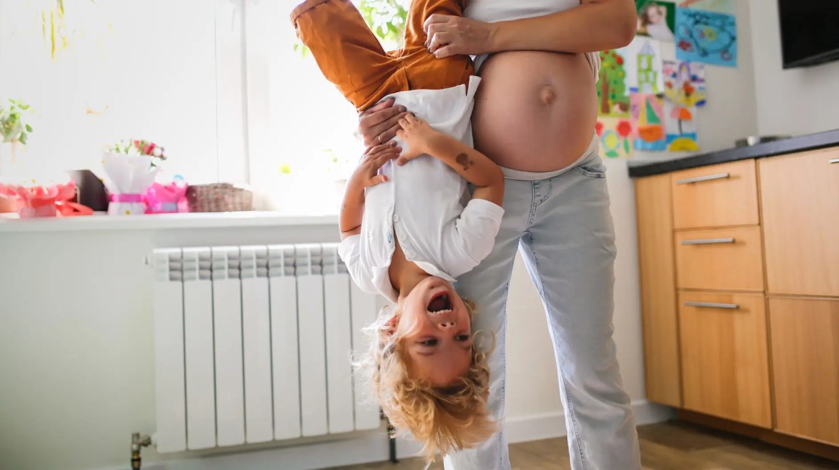 Schwangere Frau hebt ihr erstes Kind an den Füßen nach oben, das lacht herzlich dabei.