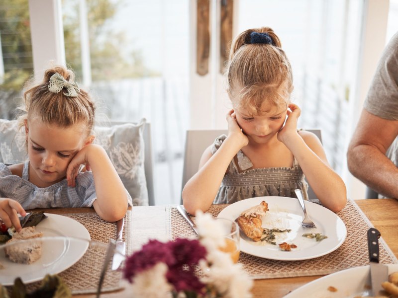 Zwei Mädchen sitzen am Familientisch und schauen das Essen auf ihren Tellern skeptisch an.