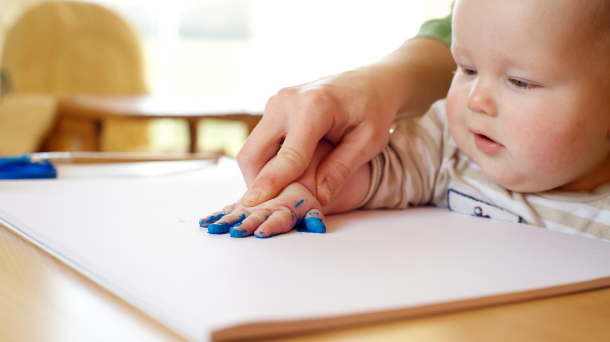 Ein Kleinkind macht mit Mamas Hilfe und Fingerfarbe einen Handabdruck auf einem Blatt Papier