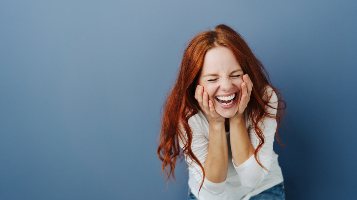 Frau hockt vor einem blauen Hintergrund mit roten Haaren und lacht