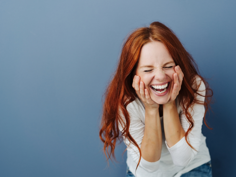 Frau hockt vor einem blauen Hintergrund mit roten Haaren und lacht