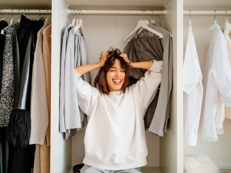 Frau, die in ihrem vollen Kleiderschrank sitzt und die Zunge rausstreckt.