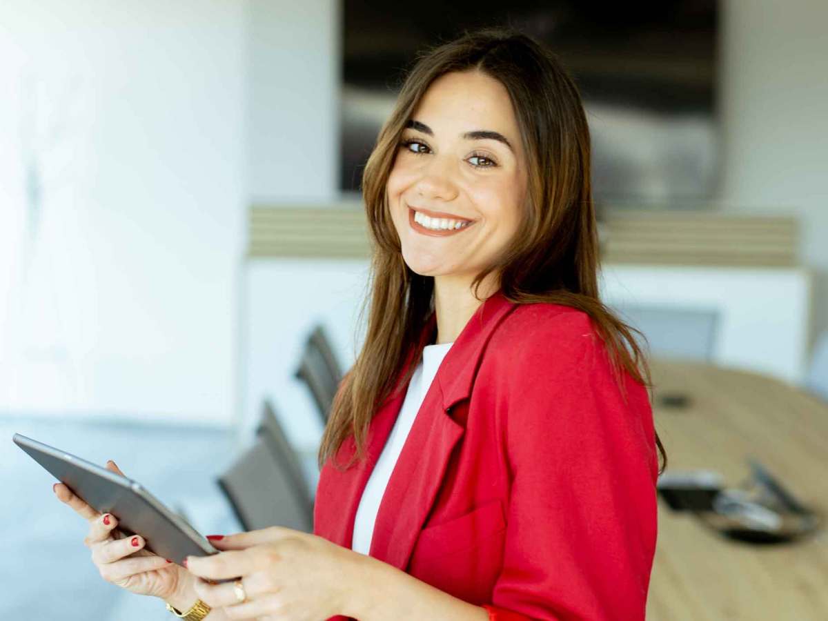Junge Frau mit rotem Blazer steht im B&uuml;ro und h&auml;lt ein Tablet fest.