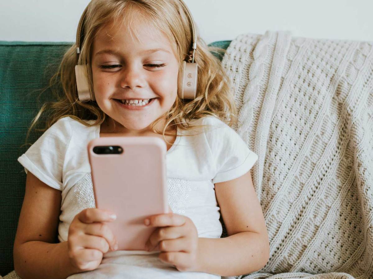 M&auml;dchen, ca. 6 Jahre, sitzt mit Kopfh&ouml;rern und Handy auf dem Sofa und schaut sich etwas auf dem kleinen Bildschirm an.