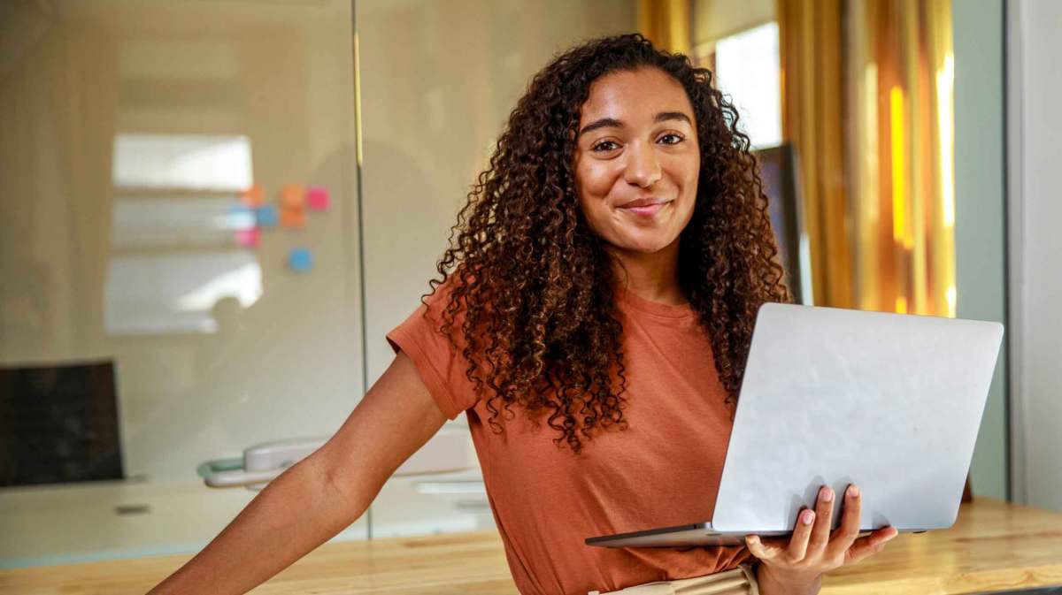 Junge Frau steht im Büro angelehnt und hält einen Laptop in der Hand.