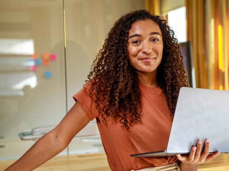 Junge Frau steht im Büro angelehnt und hält einen Laptop in der Hand.