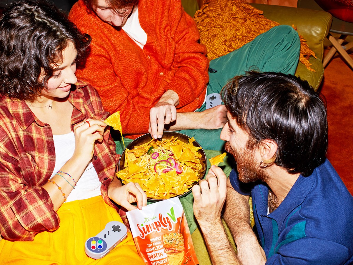 Drei junge Freunde in bunter Kleidung sitzen auf einem Sofa und teilen eine Schüssel Nachos mit Käse und Zwiebeln.