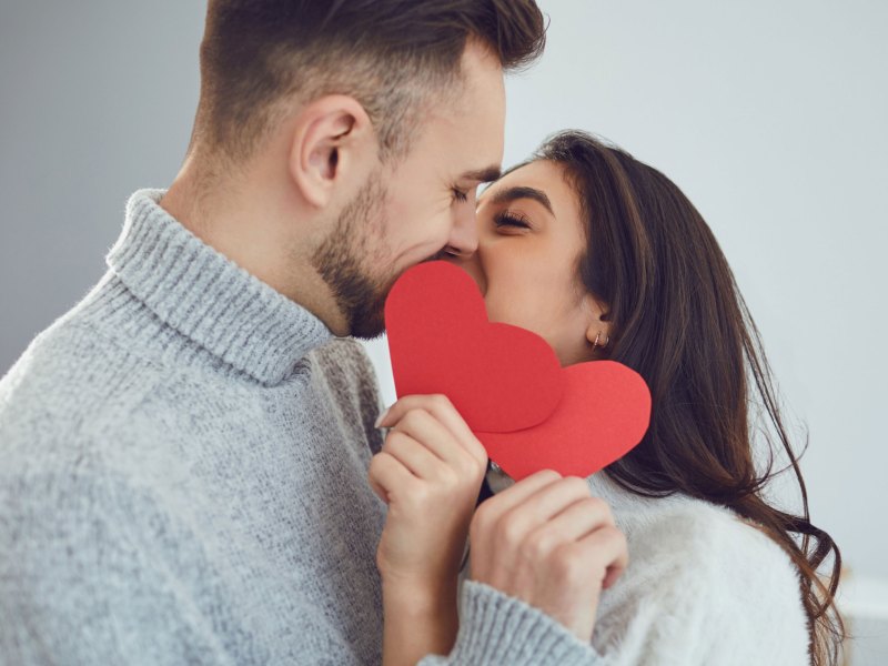 Frau und Mann küssen sich und halten sich rote Papierherzen vor das Gesicht