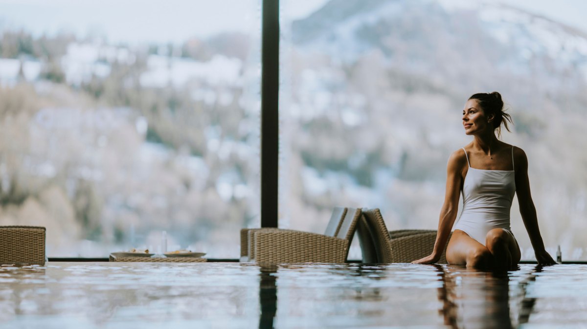 Frau sitzt auf Beckenrand eines Schwimmbeckens, in einem Wellnesshotel in den verschneiten Bergen.
