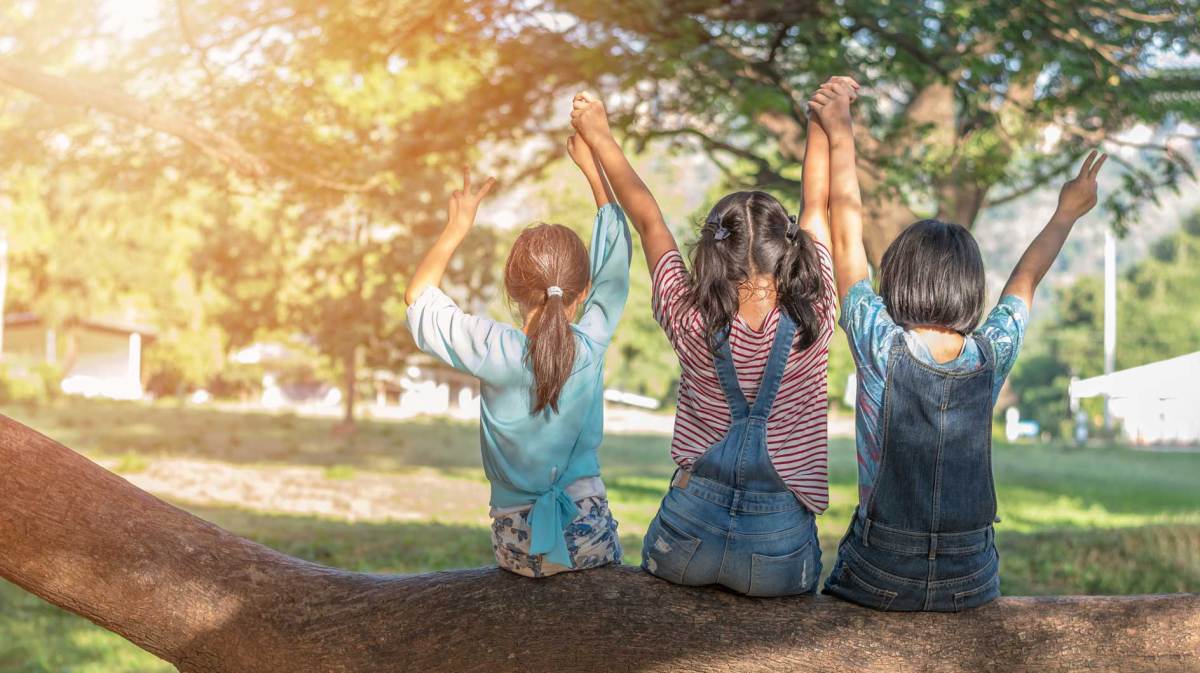 Drei Mädchen sitzen auf einem dicken Ast eines Baumes, haben ihre Hände in die Höhe gestreckt, halten einander fest und zeigen das Peace-Symbol.