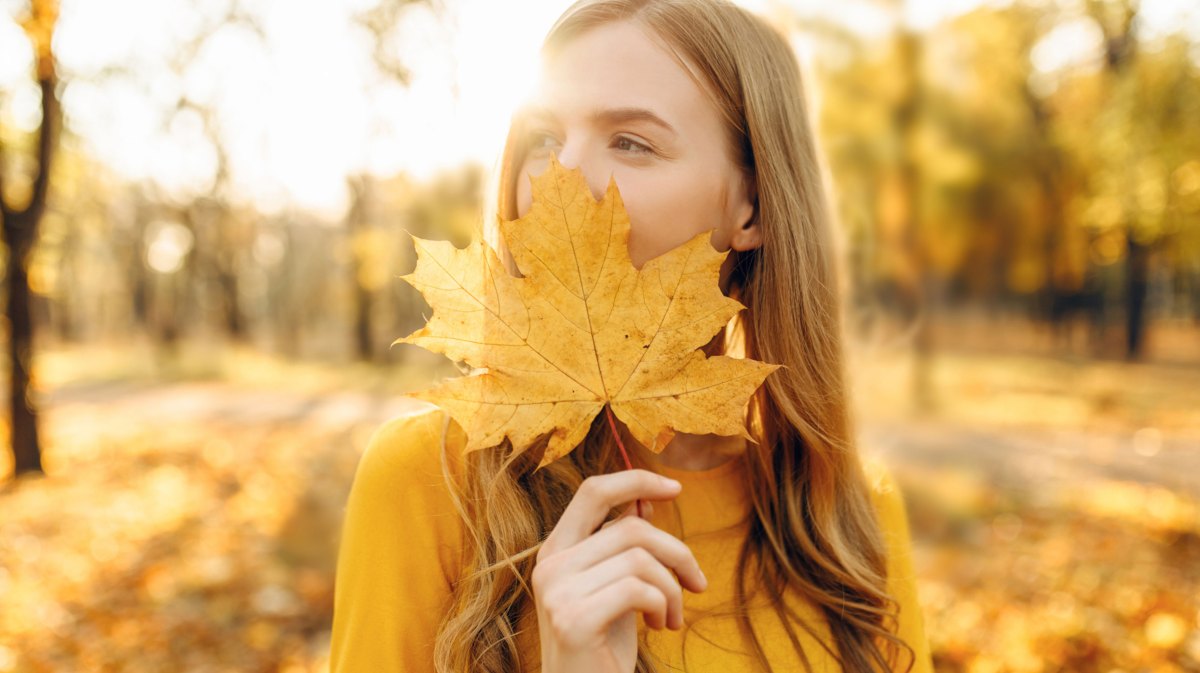 Frau im herbstlichen Wald hält sich Ahornblatt vor das Gesicht
