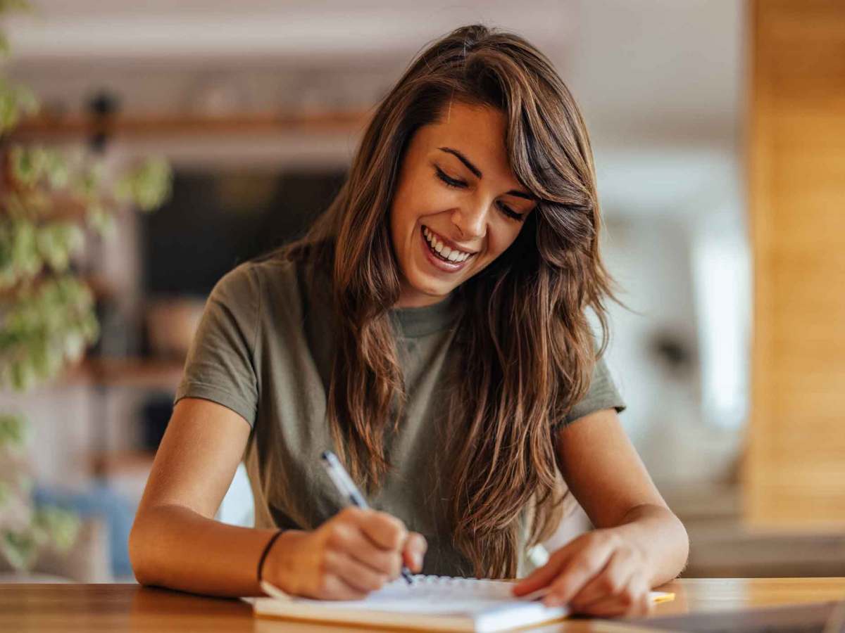 Frau sitzt an einem Schreibtisch und schreibt etwas auf und lacht dabei.