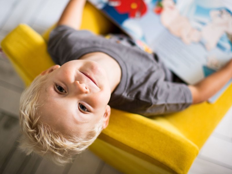Junge, ca. 5 Jahre, sitzt auf einem gelben Kindersessel, hat ein Buch auf dem Scho&szlig; und schaut nach oben in die Kamera.