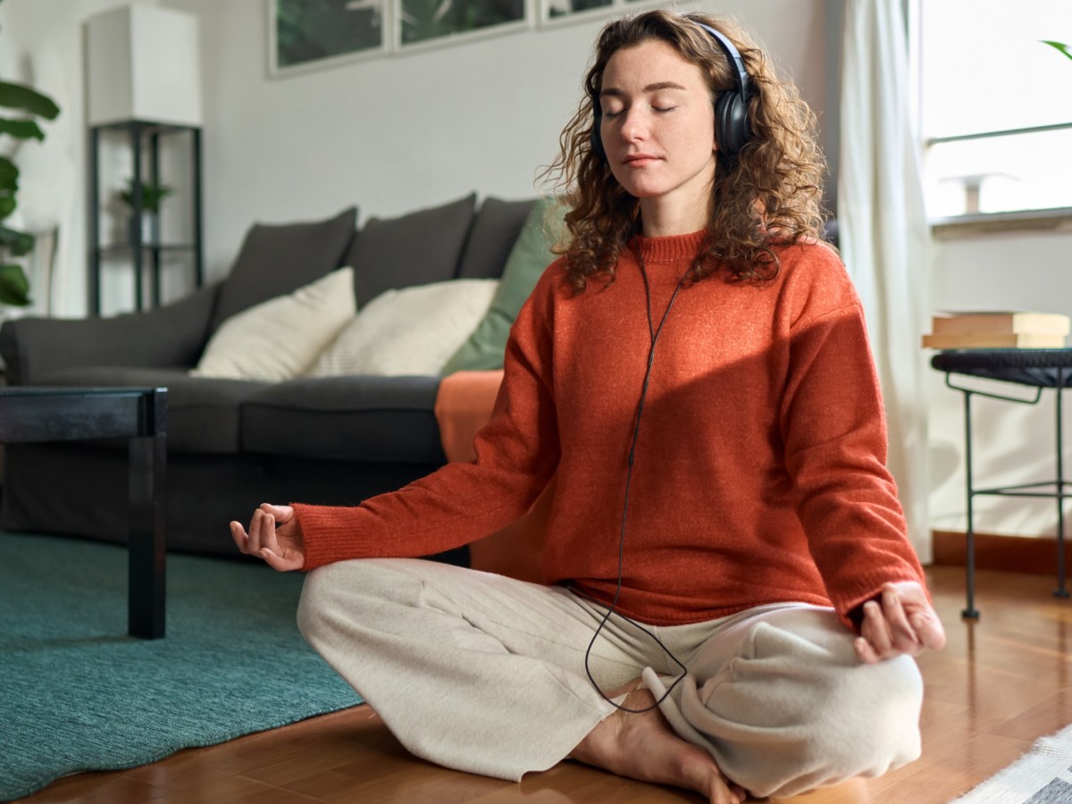 Frau meditiert im Wohnzimmer und hat dabei Kopfh&ouml;rer auf.