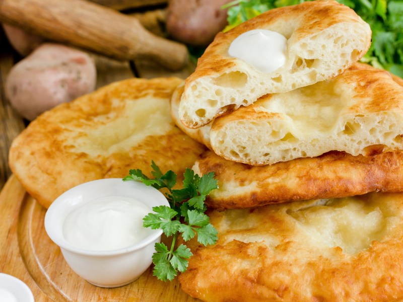 Mehrere Langosch, gestapelt auf Holzbrett, daneben ein Schüsselchen mit Saurer Sahne.