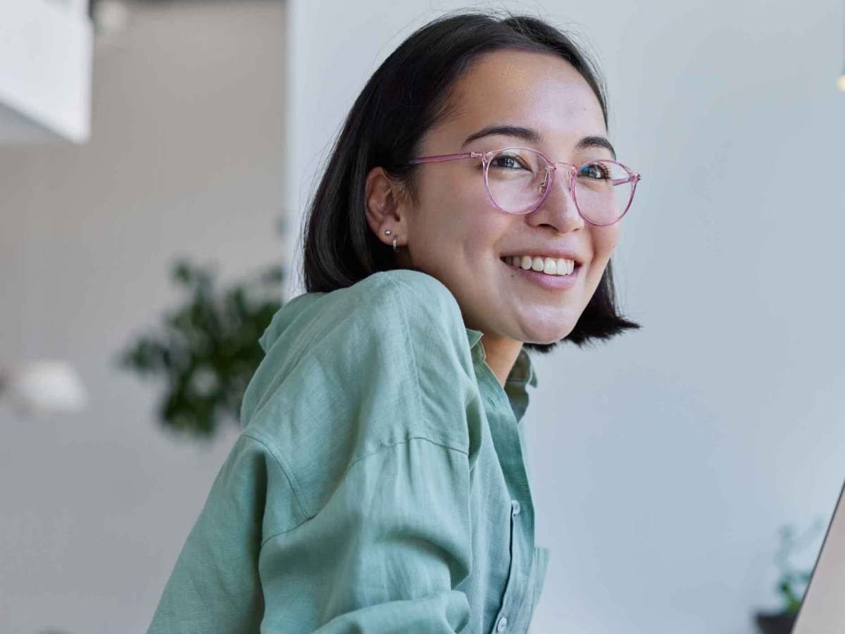 Dunkelhaarige Frau mit Brille und einer salbei-farbenen Bluse sitzt am Laptop.