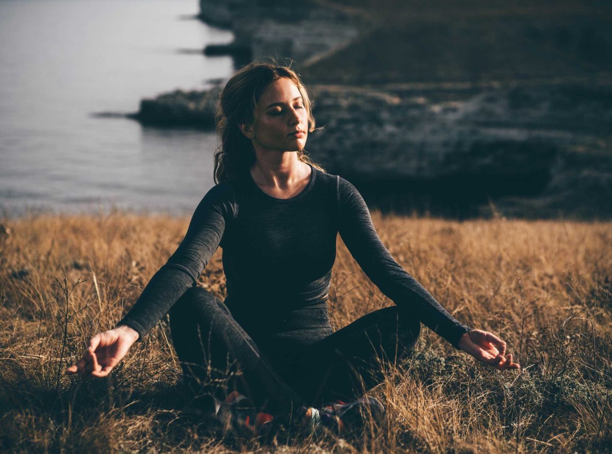 Junge Frau in schwarzem Hemd übt Yoga-Asanas auf hoher Felsenküste über dem Meer bei hellem Sonnenlicht und leichtem Wind in ruhigem Morgen