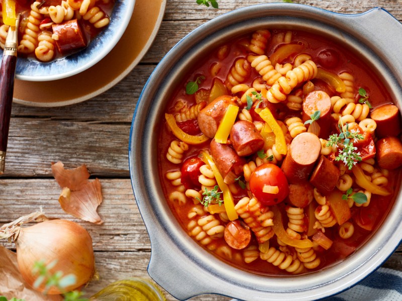 Wurstgulasch mit Nudeln und Paprika in großem Topf auf Holztisch.