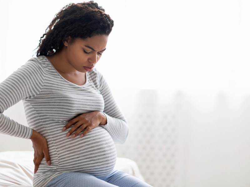 Schwangere Frau sitzt mit Wehen auf dem Bett zu Hause und hat eine Hand jeweils auf Bauch und Rücken gelegt.