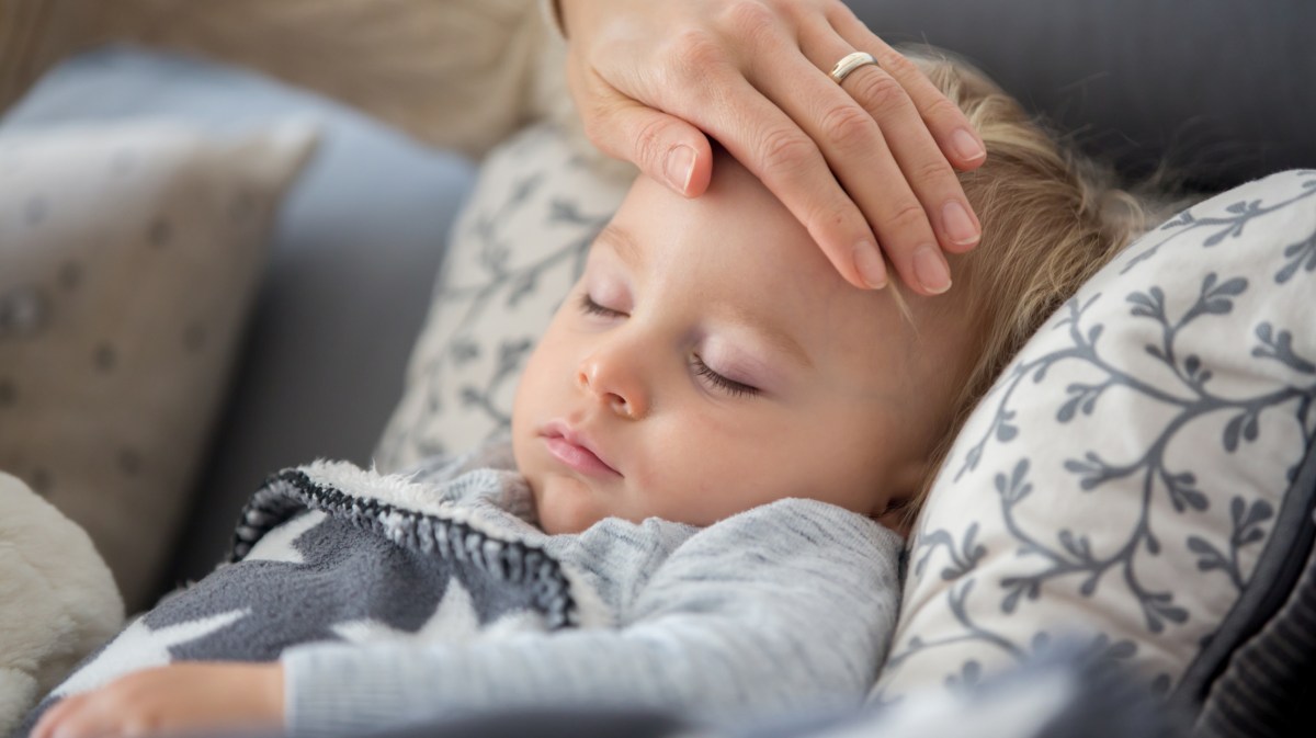 Krankes KInd liegt auf dem Sofa. Die Hand der Mutter fühlt den Kopf.