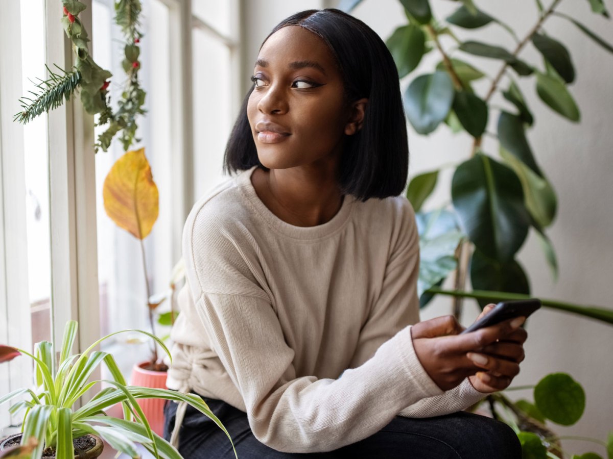 Frau sitzt am Fenster, das Handy in der Hand und schaut nachdenklich glücklich nach draußen.