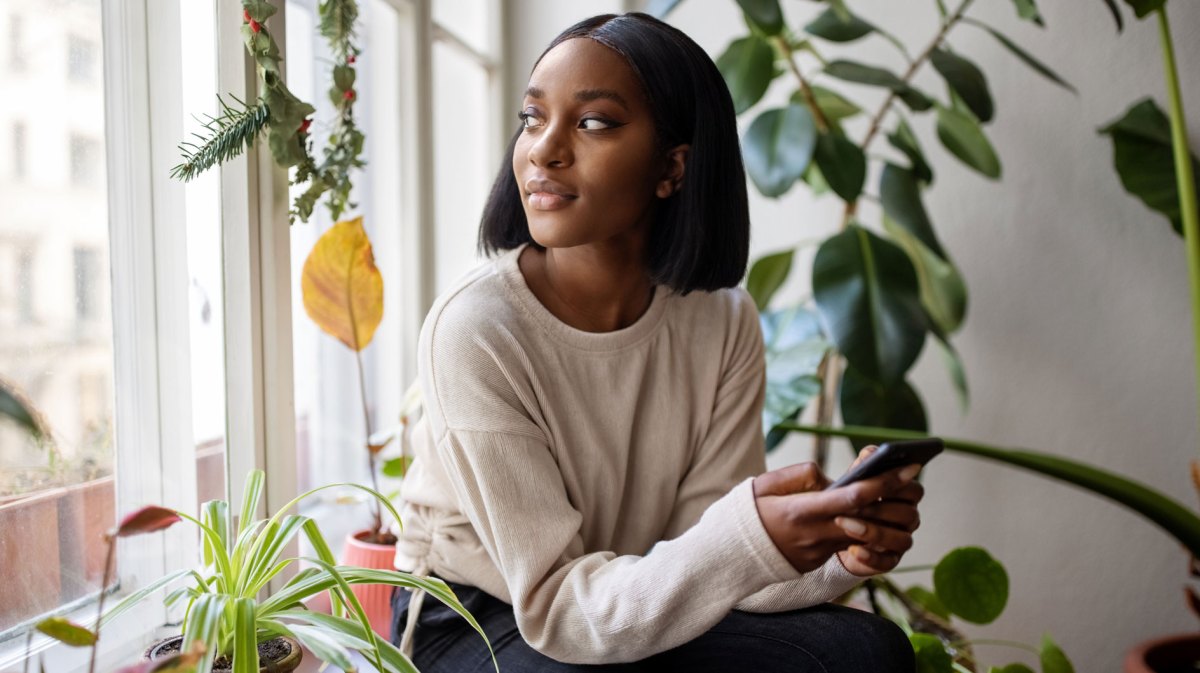 Frau sitzt am Fenster, das Handy in der Hand und schaut nachdenklich glücklich nach draußen.