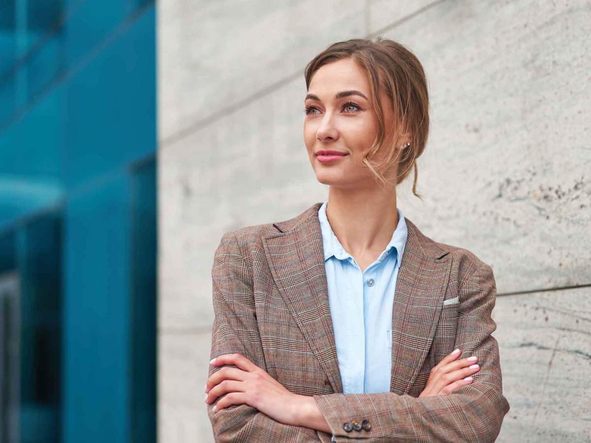 Frau mit Blazer steht mit verschr&auml;nkten Armen vor einem B&uuml;rogeb&auml;ude.