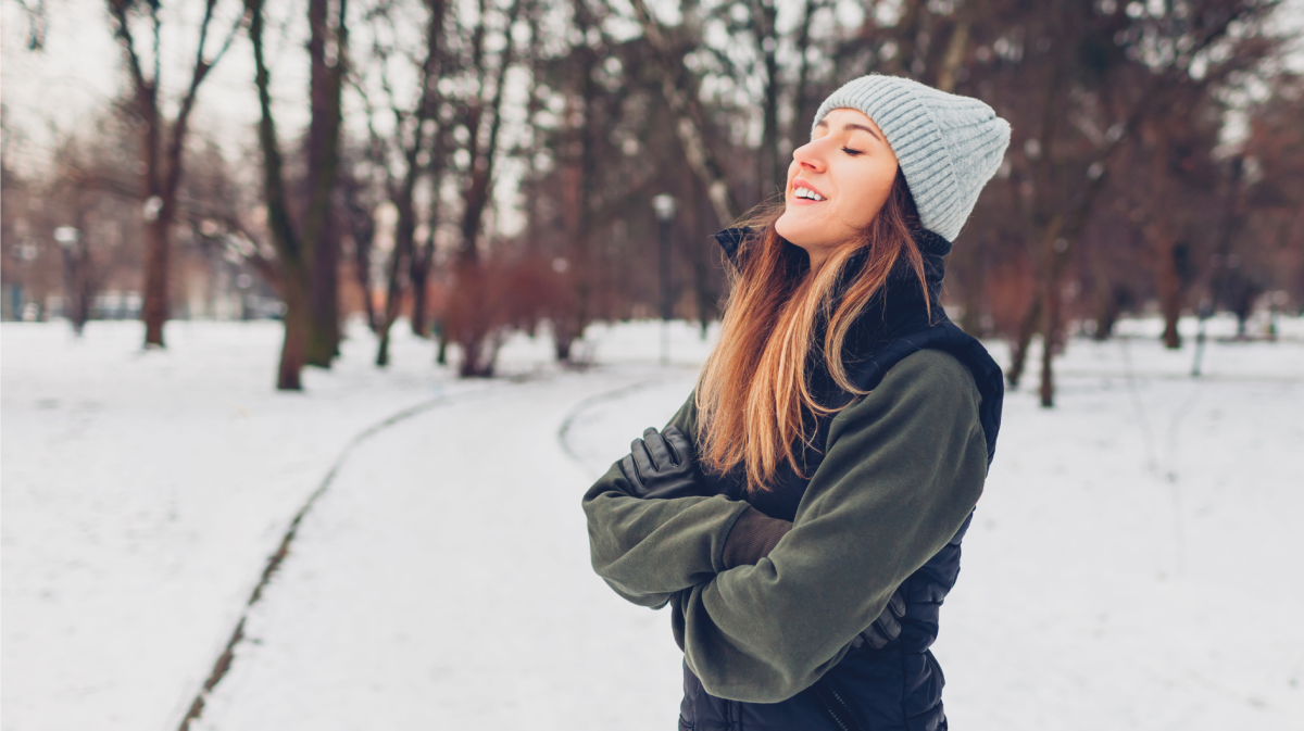 Junge Frau mit langen braunen Haaren und grauer Strickmütze steht lächelnd in einem verschneiten Park und genießt die Winterluft.