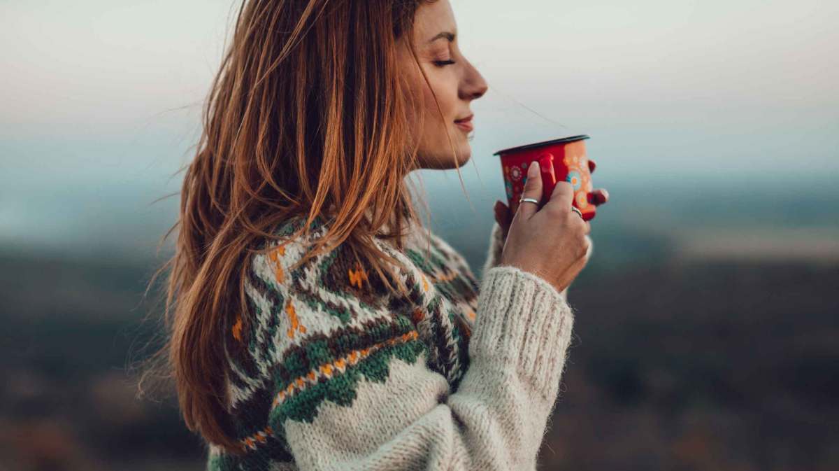 Frau mit Tasse und Strickpullover steht draußen vor einer Landschaft.