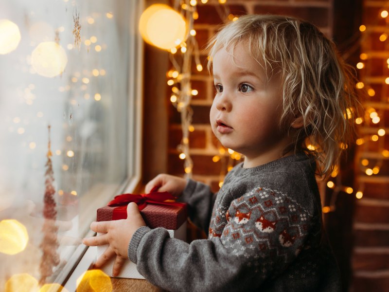 Kleinkind schaut neugierig und gebannt aus dem Fenster. Im Hintergrund leuchten kleine Lampen.
