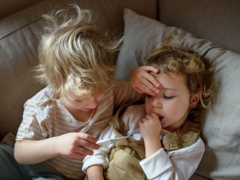Zwei Kinder liegen im Bett. Der große Bruder, ca. 5 Jahre, legt seiner kleinen Schwester, ca. 3 Jahre, die Hand auf die Stirn und schaut auf das Fieberthermometer in seiner Hand.