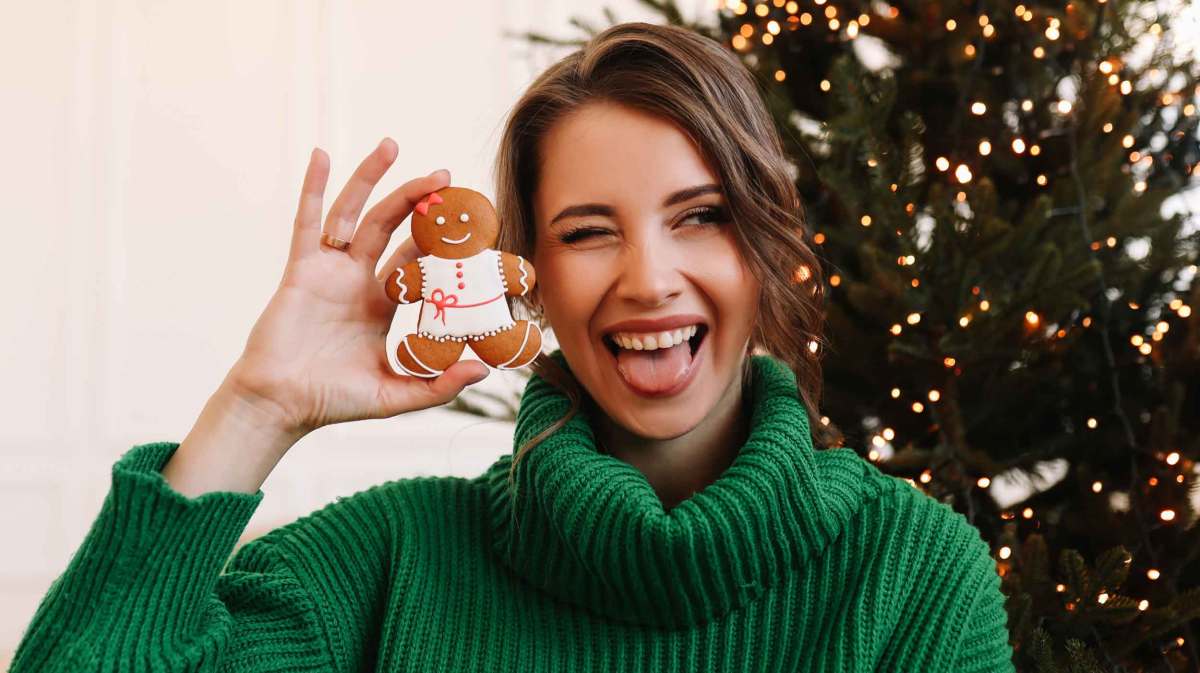 Frau mit grünem Pullover hält einen Lebkuchenmann in der Hand und zwinkert dabei. Im Hintergrund steht ein Weihnachtsbaum.