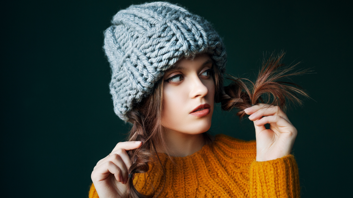 Eine junge Frau in einem gelben Strickpullover und einer grob gestrickten grauen Mütze. Sie hält spielerisch ihre braunen Haarspitzen mit den Händen, während sie seitlich schaut. Der Hintergrund ist dunkelgrün.
